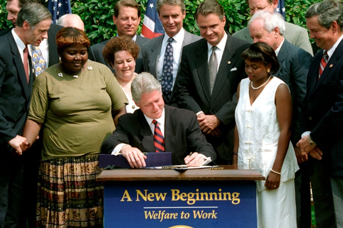 Bill Clinton is seated at a desk outside, signing a bill. Surrounding him are white men politicians, 1 white woman, and 2 Black women.
