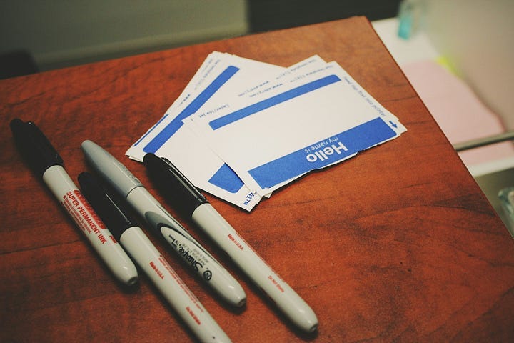 A small stack of blank sticker name tags reading “Hello, My Name Is”, next to four markers.