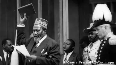 A black and white photo of Jomo Kenyatta, Kenya's first president, holding up a black book in his right and a white piece of paper in his left