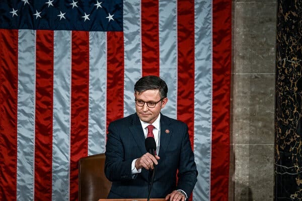 Speaker Mike Johnson adjusting a microphone. An American flag is on display behind him.