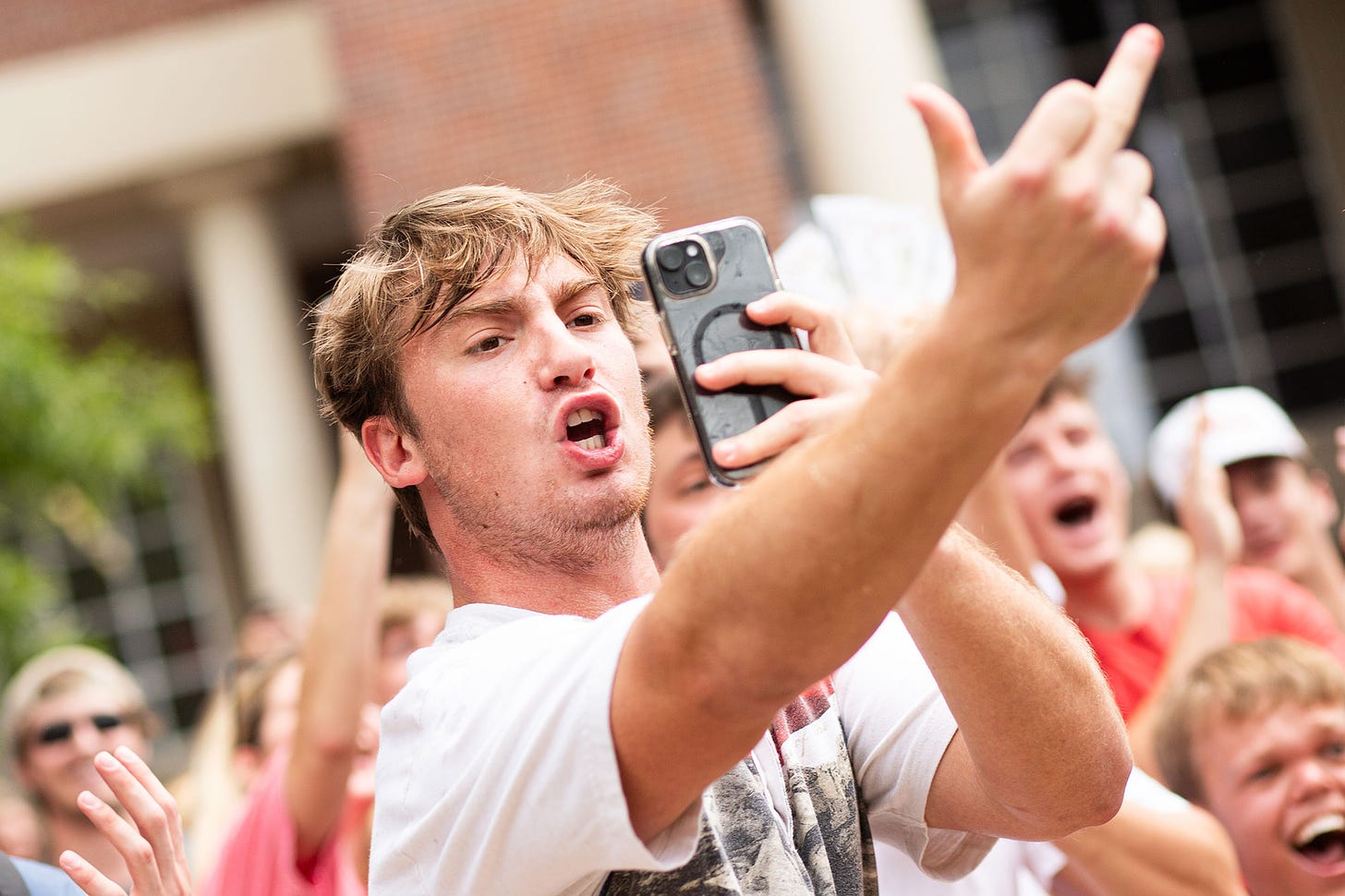 a male student holds a phone in one hand and sticks his other hand out flipping someone off