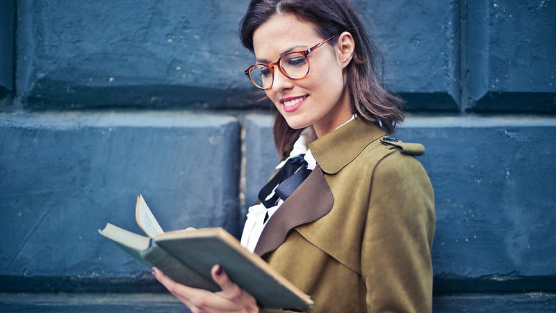 Woman in brown suede peacoat and glasses reading a book
