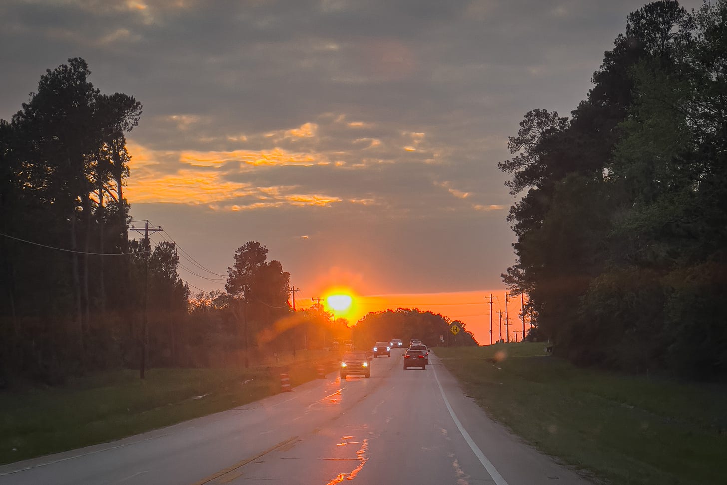 A country road with the sun-setting on the horizon;cars approaching with headlights, and the sun’s reflection on the wet asphalt road