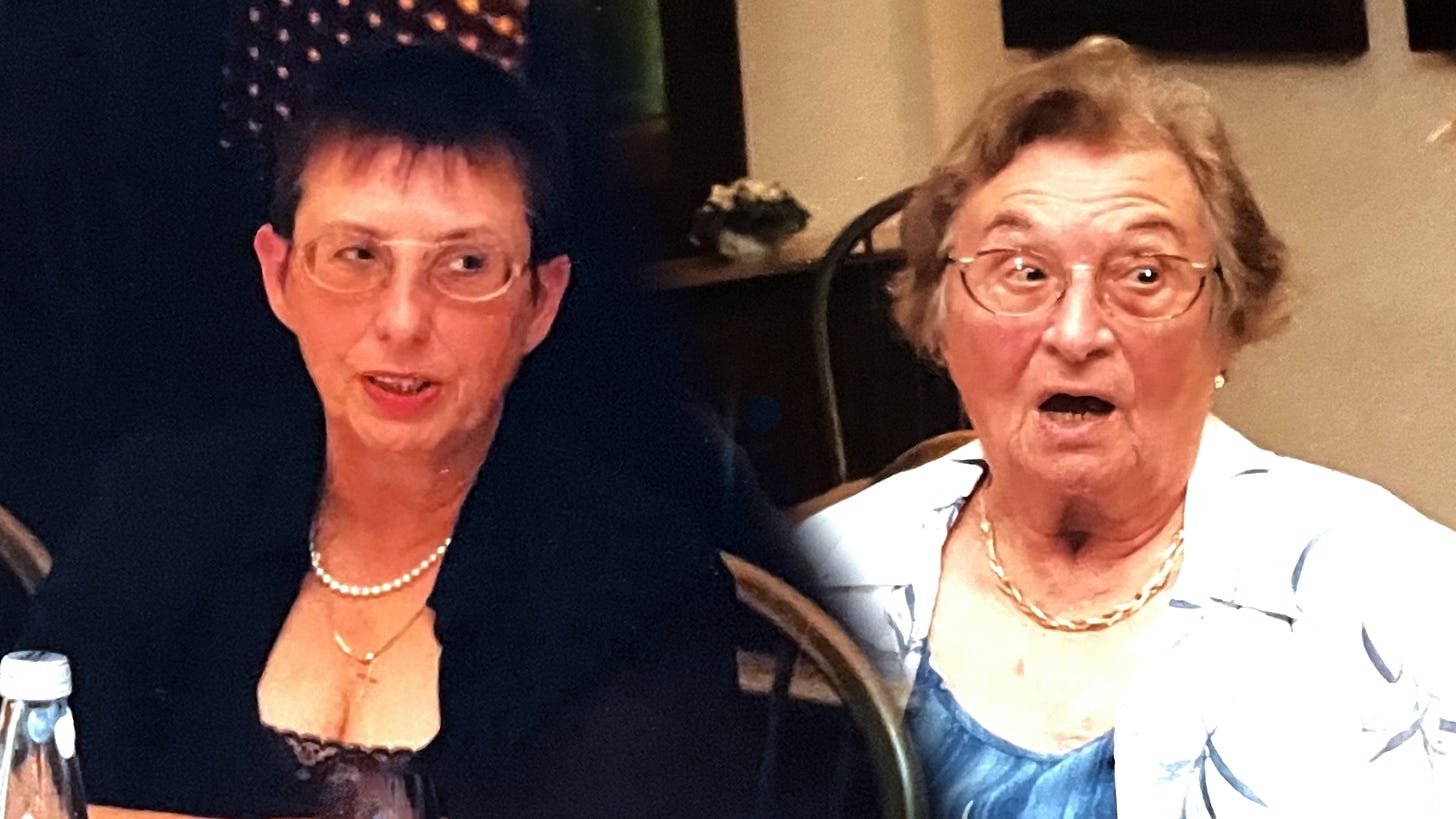 Two older women sit side by side at a formal dinner table. The woman on the left wears glasses, a black jacket over a lace-trimmed dress, and a pearl necklace; she looks mid-sentence, composed and quietly observant. The woman on the right, wearing a light blue dress and gold chain, appears caught mid-reaction—her eyes wide, mouth open in expressive surprise. Both women reflect a moment of vivid, intergenerational presence. Two older women sit side by side at a formal dinner table. The woman on the left wears glasses, a black jacket over a lace-trimmed dress, and a pearl necklace; she looks mid-sentence, composed and quietly observant. The woman on the right, wearing a light blue dress and gold chain, appears caught mid-reaction—her eyes wide, mouth open in expressive surprise. Both women reflect a moment of vivid, intergenerational presence.
