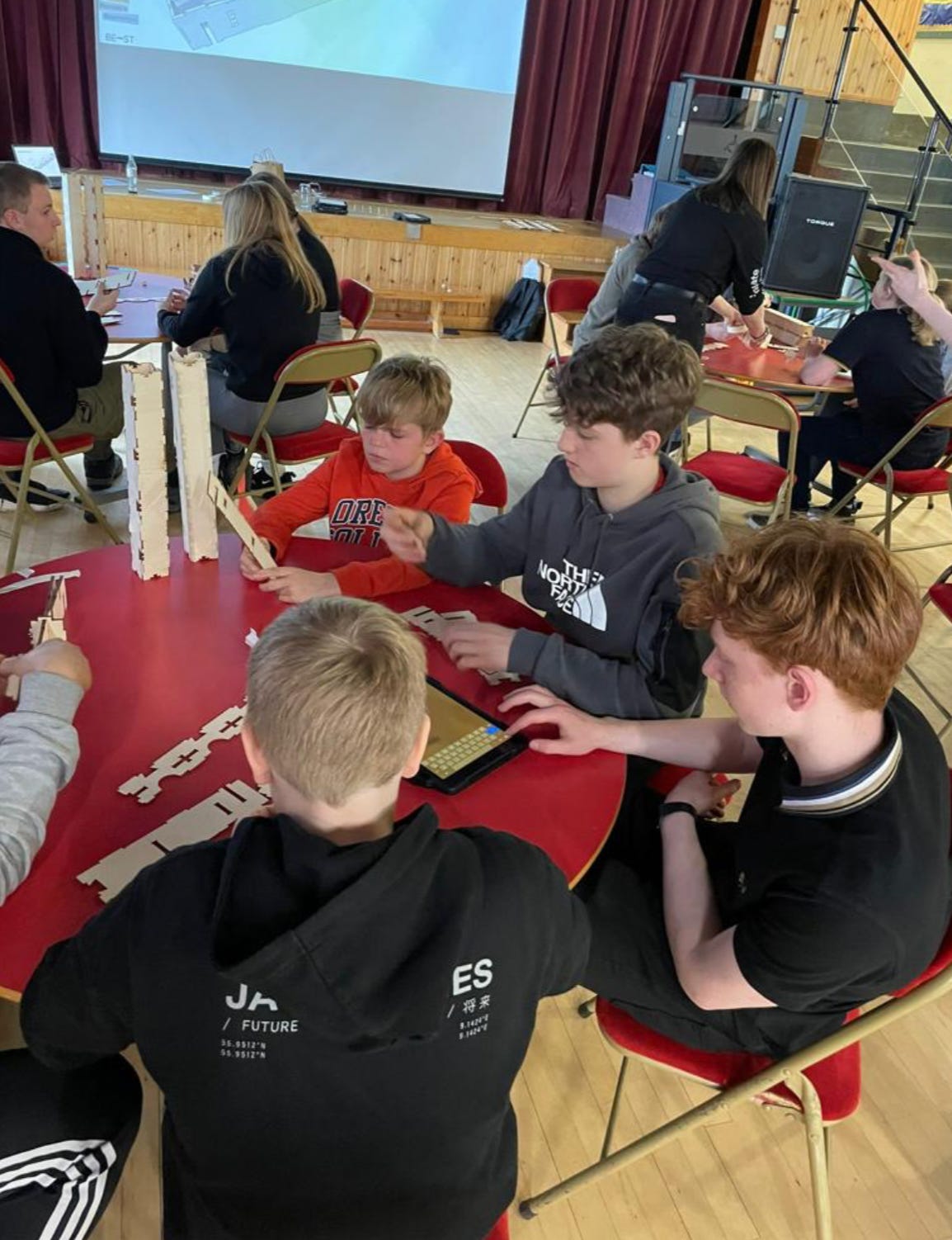 School children sit around a table assembling WikiHouse block scale models. Behind them a screen shows an assembled WikiHouse beam.