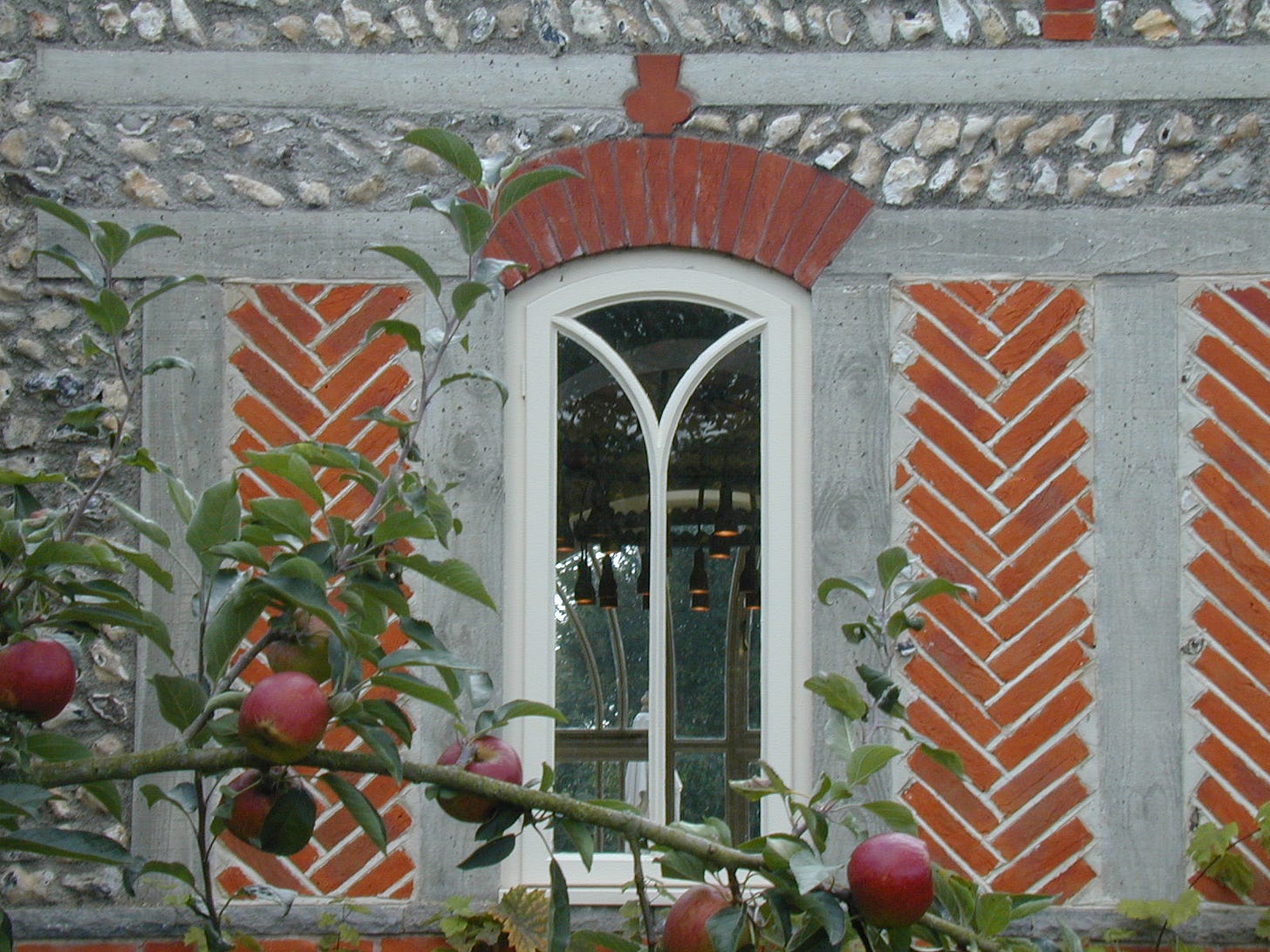 Detail of the wall of the West Dean Visitors' Centre, showing the brick and rough stone, curved window mullions, and the branch of an apple tree growing near the wall.