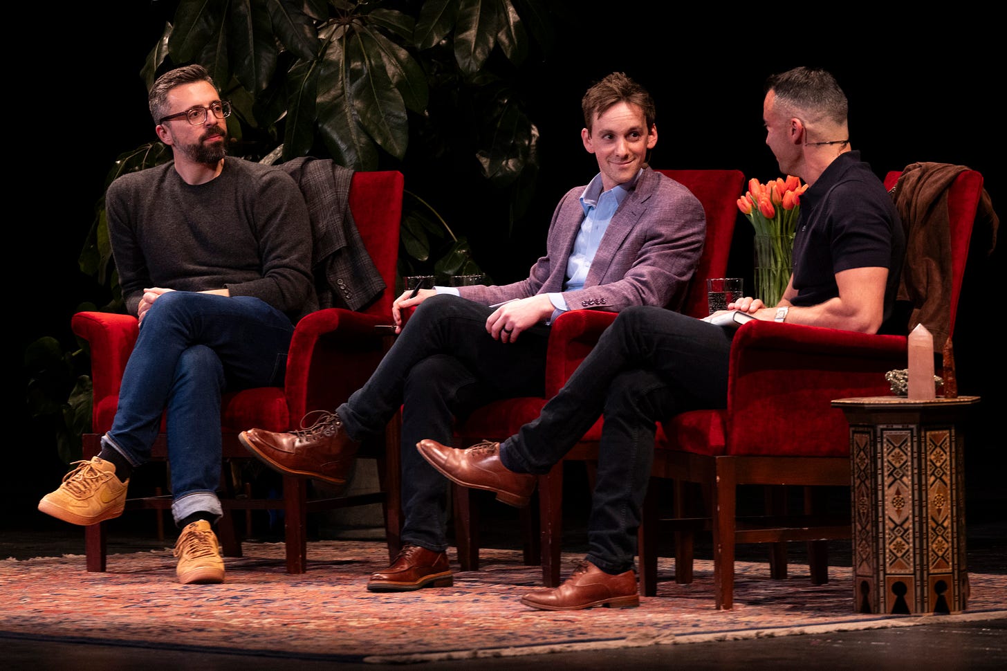 Three men sit in red chairs on stage, engaged in conversation. One holds a notebook. There are plants, tulips, and a patterned table beside them.