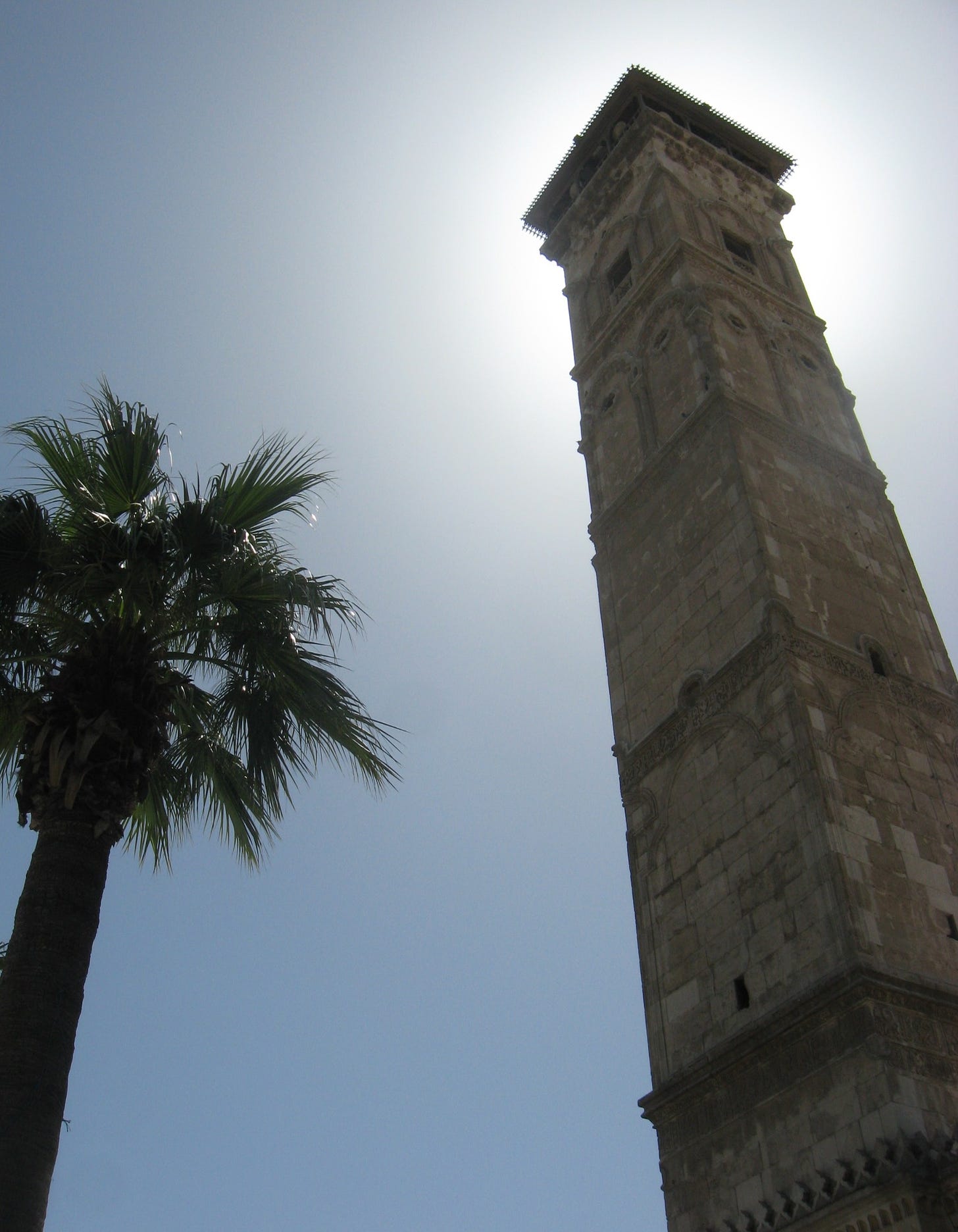 A palm tree stands beside a tall white stone minaret under a cloudless blue sky