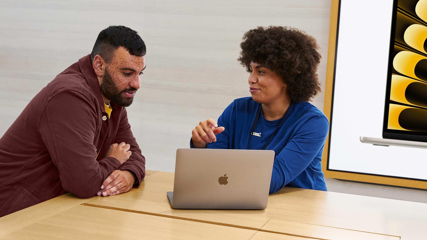 A hero image from the Today at Apple homepage. A store employee sits behind a MacBook and explains content onscreen to a customer.