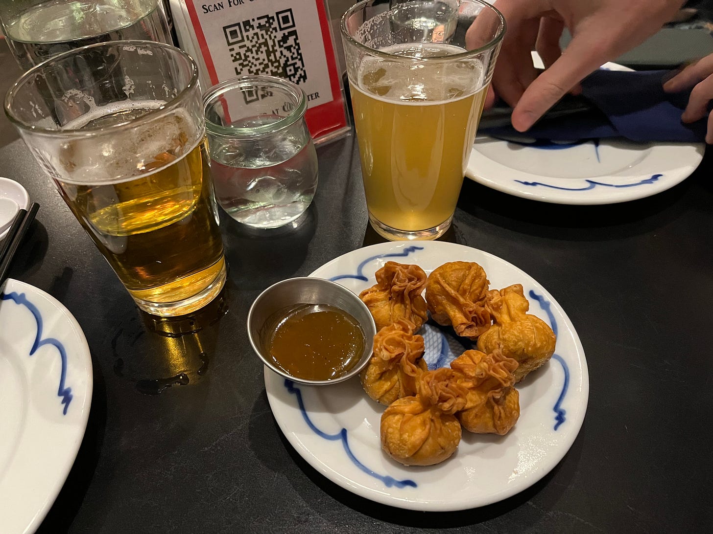 On a black table, there's a white porcelain plate with six golden, deep-fried dumplings. The dumpling wrappers are crisp and bubbly. Also on the plate there's a dish of dipping sauce, which is brown. There are cups of beer and water on either side of this plate.