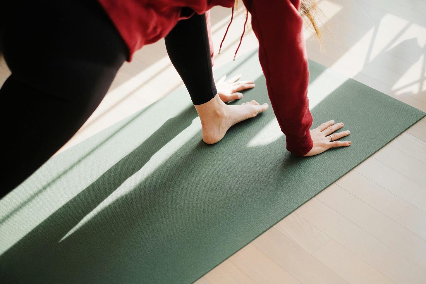 A person in red sweatshirt and black leggings in a forward lunge on a yoga mat, close-up view so only hands and front foot and part of trunk are visible