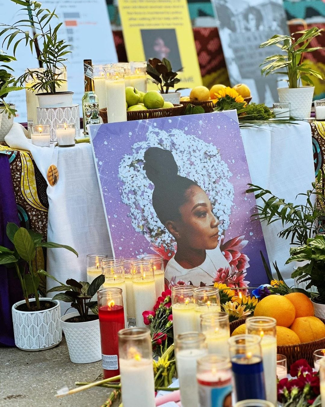 A photos of a memorial altar. At the center, leaning on a table surrounded by dozens of candles and plants in white pots, is a digital illustration of a Black woman with a high bun surrounded by white and red flowers.