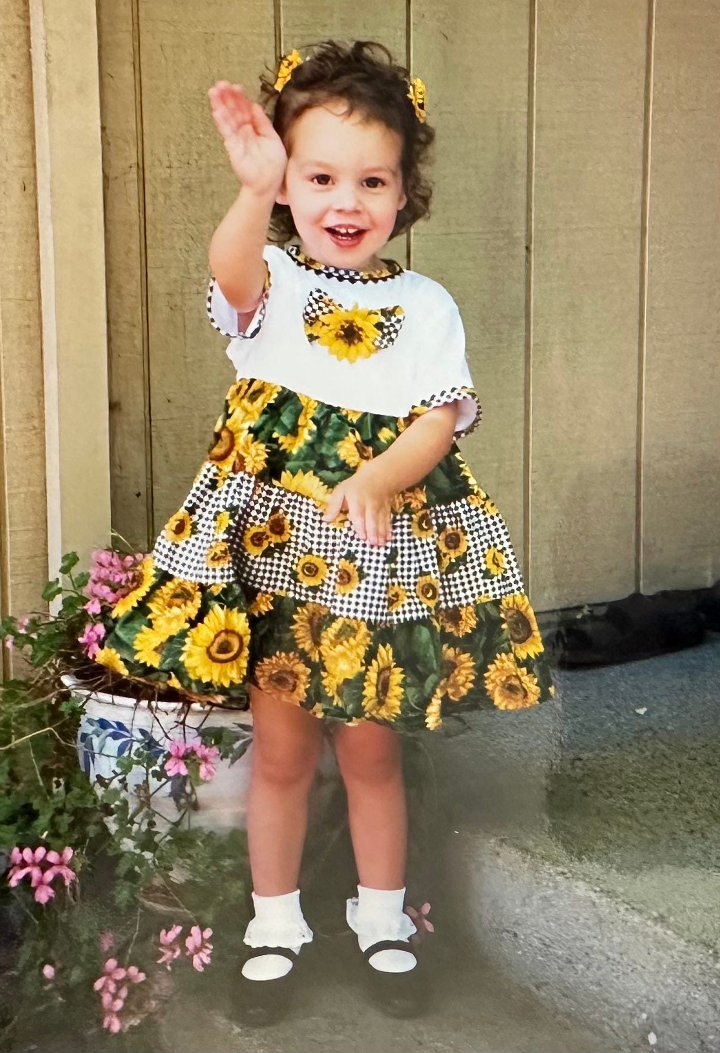 2 year old Chloe waves at the camera with an easy smile. her dress is alternating fabric of sunflowers and black and white checkered print, and she has sunflower clips in her hair.