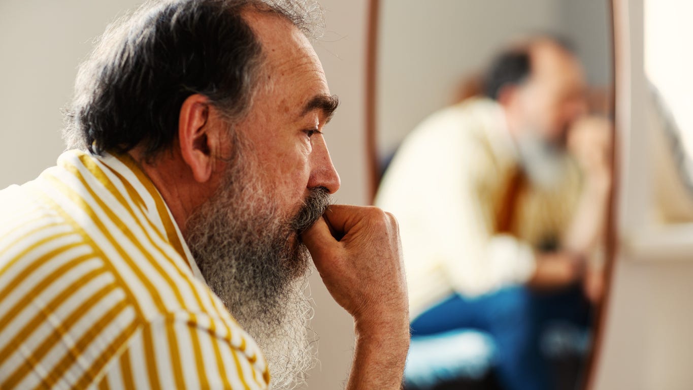 Aging white man with graying beard in thoughtful profile pose is blurredly reflected in mirror next to him. His forefinger and thumb are nestled into his upper lip.