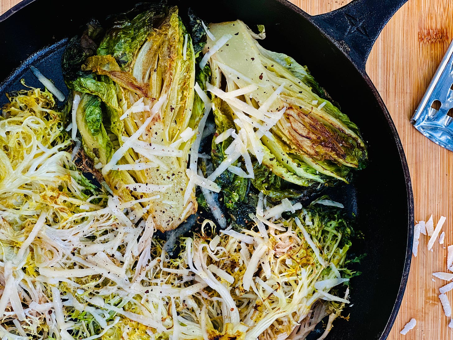 Pan-seared baby lettuce heads (gem and frisee) in a cast-iron skillet with shredded parmesan and black pepper over top