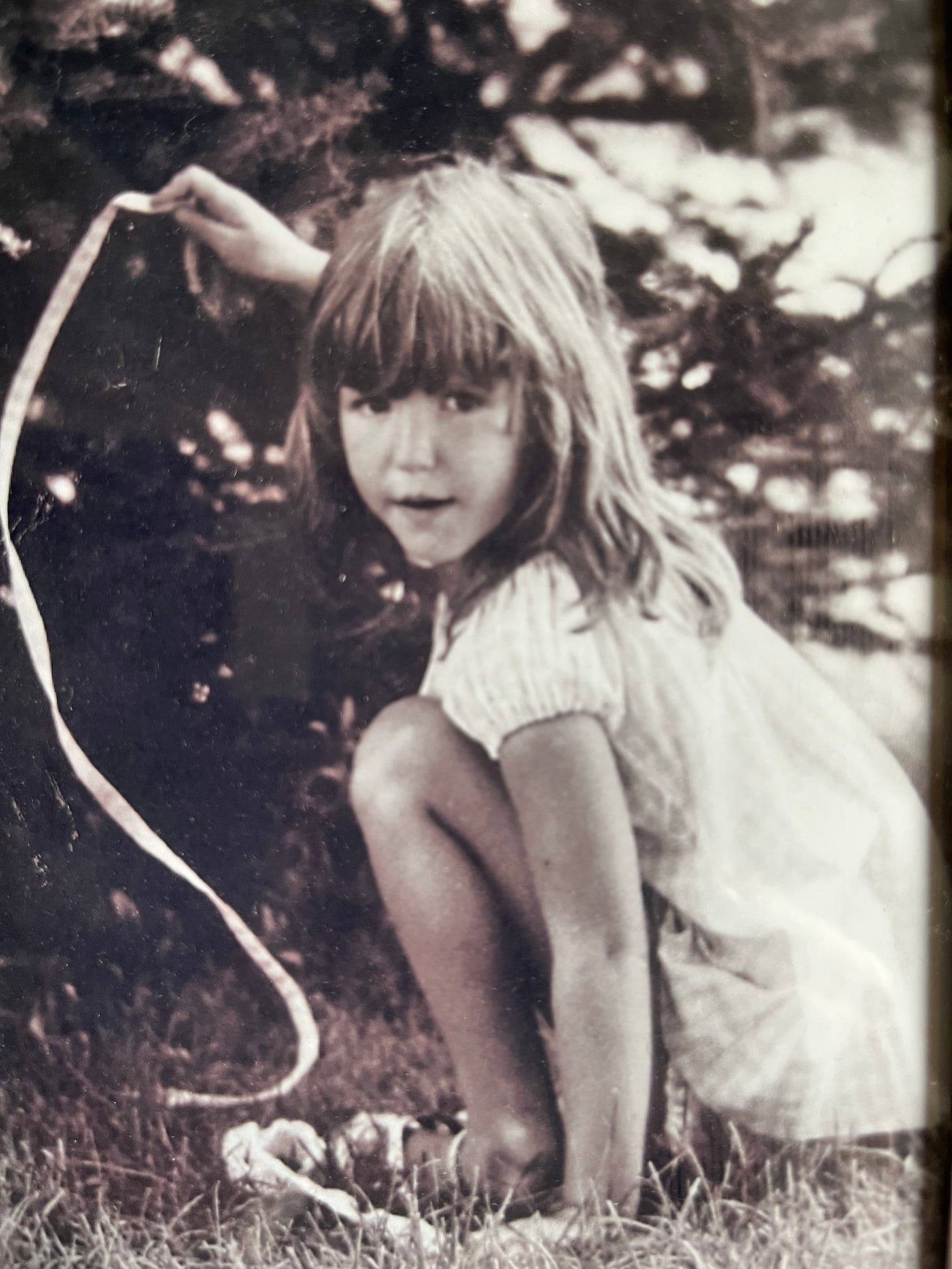 A black and white photograph of Tracy Livecchi as a child, crouching in the grass and wearing a dress.