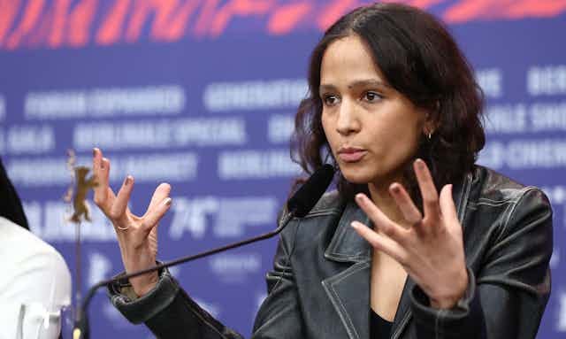 A woman in a denim jacket gestures with both hands, fingers splayed, as she talks into a microphone at a desk.