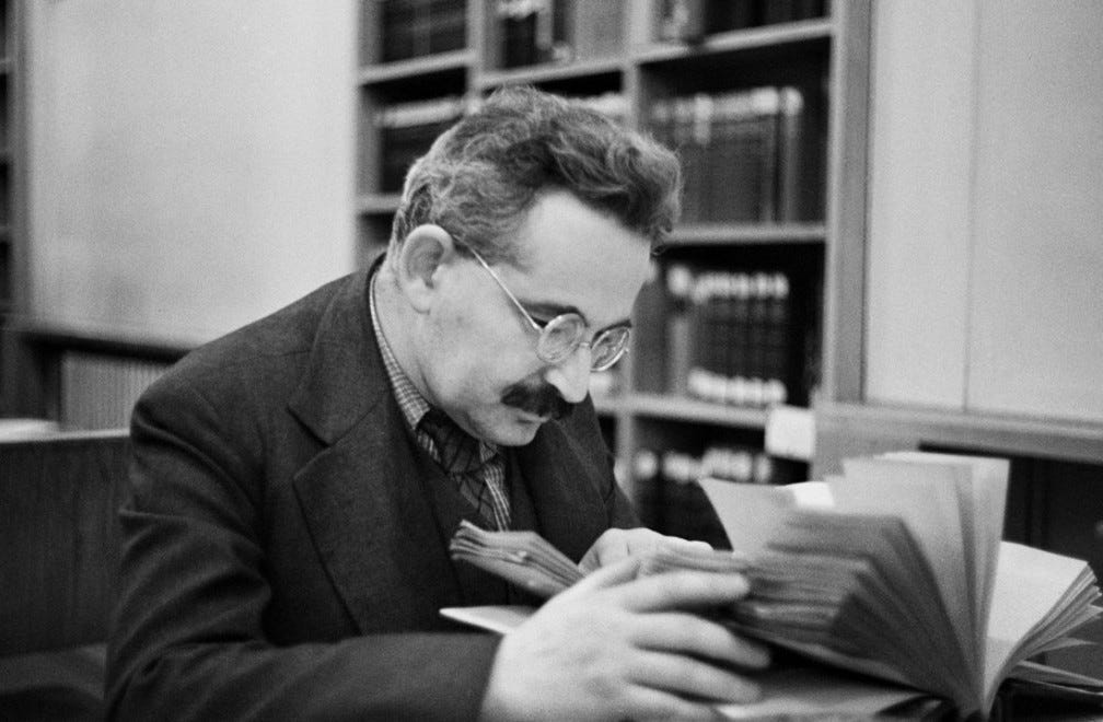 A vintage photograph of Benjamin with a mustach and round glasses, bent over two open books. Modern, uniform stacks wall the room where he sits at a table.