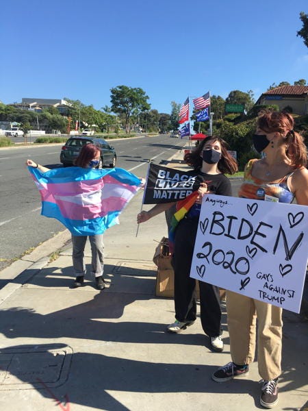 Photo of three teenage girls wearing face masks at a street corner. One holds up a trans pride flag, one a Black Lives Matter smaller flag, and one a handmade cardboard sign that says Biden 2020 and Gays Against Trump