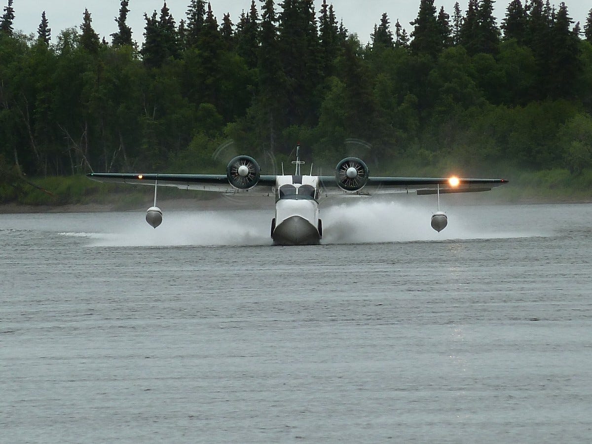 Flying boat taking off, headed straight toward viewer, with forest in the background.