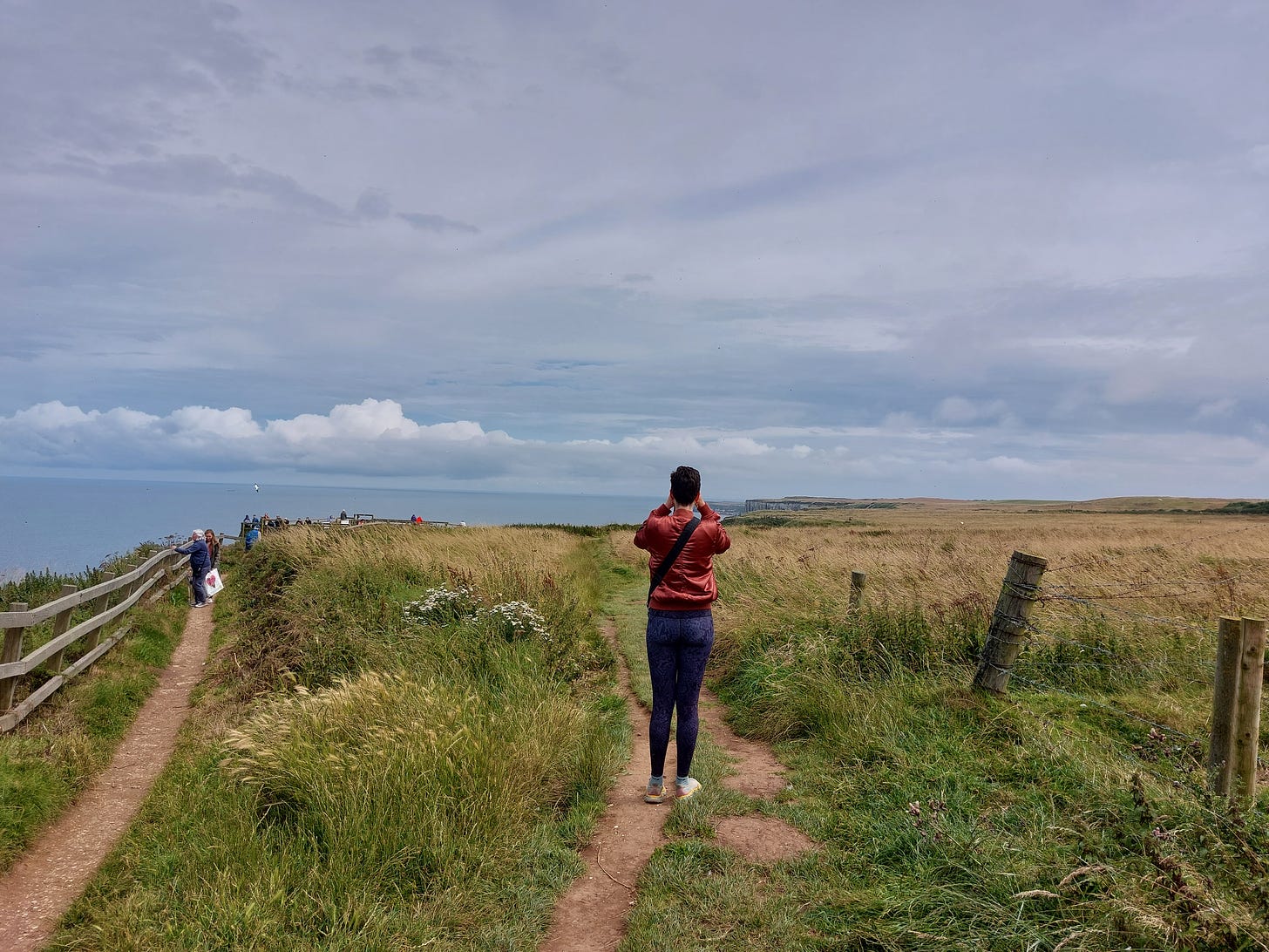 A wide shot taken from behind a white lady with short brown hair. She is standing holding her hands to her face because she's looking through binoculars at Bempton Cliffs. The sky is grey-blue and the sea is just visible. The terrain is straw coloured grass