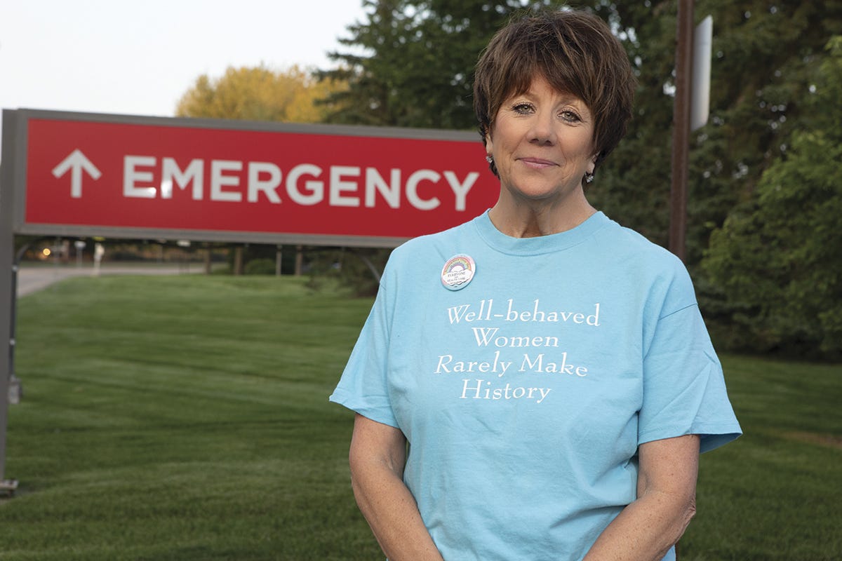 A brown haired white woman wearing a light blue shirt reading "well-behaved women rarely make history" stands in front of a red emergency hospital sign