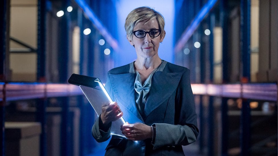 This image shows the Kerb!am Corporation's "Head of People," Judy Maddox (played by Julie Hesmondhalgh), standing in a Kerb!am warehouse corridor, looking directly into the camera, and holding a lighted clipboard (in "Kerblam!," Doctor Who Series 11's seventh episode). This image shows the Kerb!am Corporation's "Head of People," Judy Maddox (played by Julie Hesmondhalgh), standing in a Kerb!am warehouse corridor, looking directly into the camera, and holding a lighted clipboard (in "Kerblam!," Doctor Who Series 11's seventh episode).