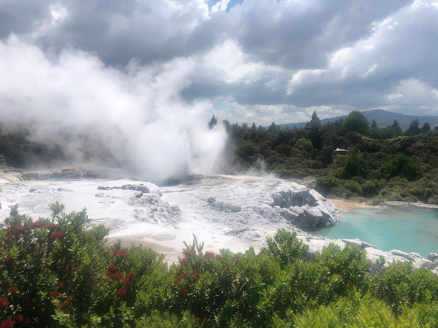 Dark clouds over a woodland landscape, with a white shelf of rock running across the middle. Water is spewing skyward from the rocks, and steam is blowing over to the left.