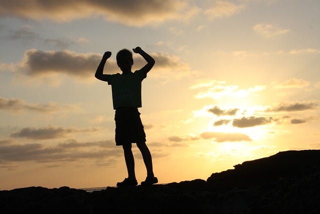 Young boy raising arms in triumph on mountain at setting of sun