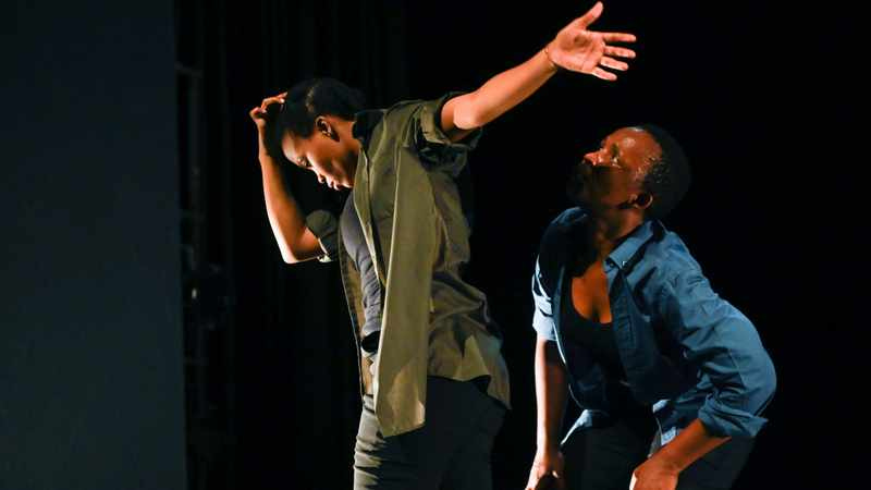 Two Black dancers, mid-performance. One is looking down, holding an arm outstretched behind. Another is just behind, looking over the other’s shoulder from a squat.