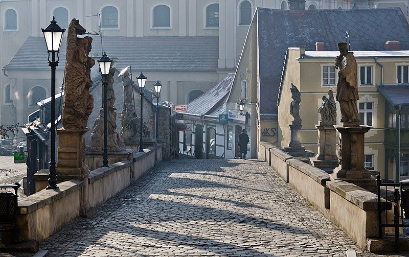  Image Description: Cobblestone bridge lined with statues, with an assemblage of ancient-looking buildings and then a looming church on the far side.