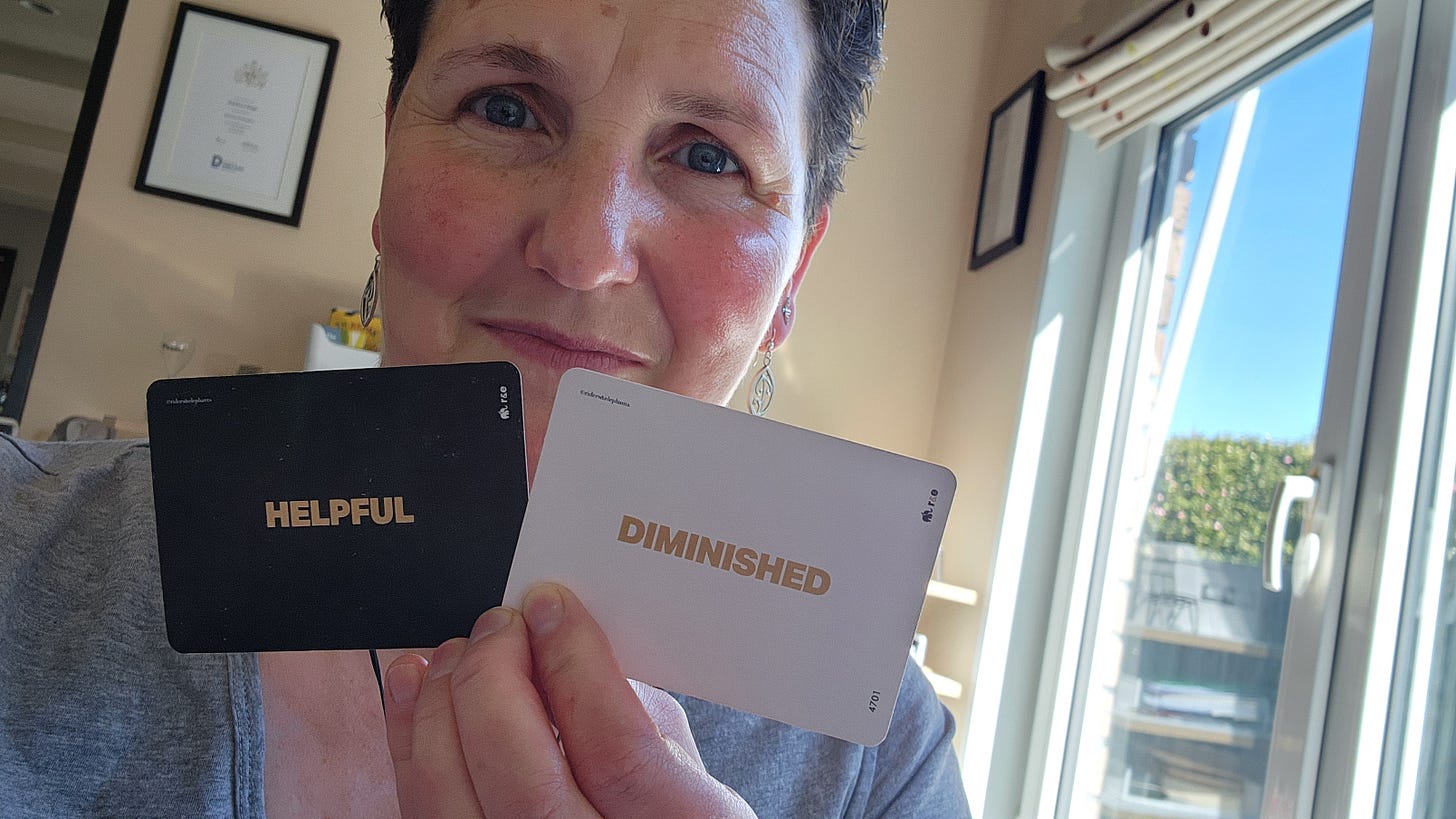 Kathryn has short brown hair and is wearing a grey marl T-shirt and holding two cards: a black card with HELPFUL and a white card with DIMINISHED. A window is visible to the right and there are a couple of framed pictures on the wall behind.