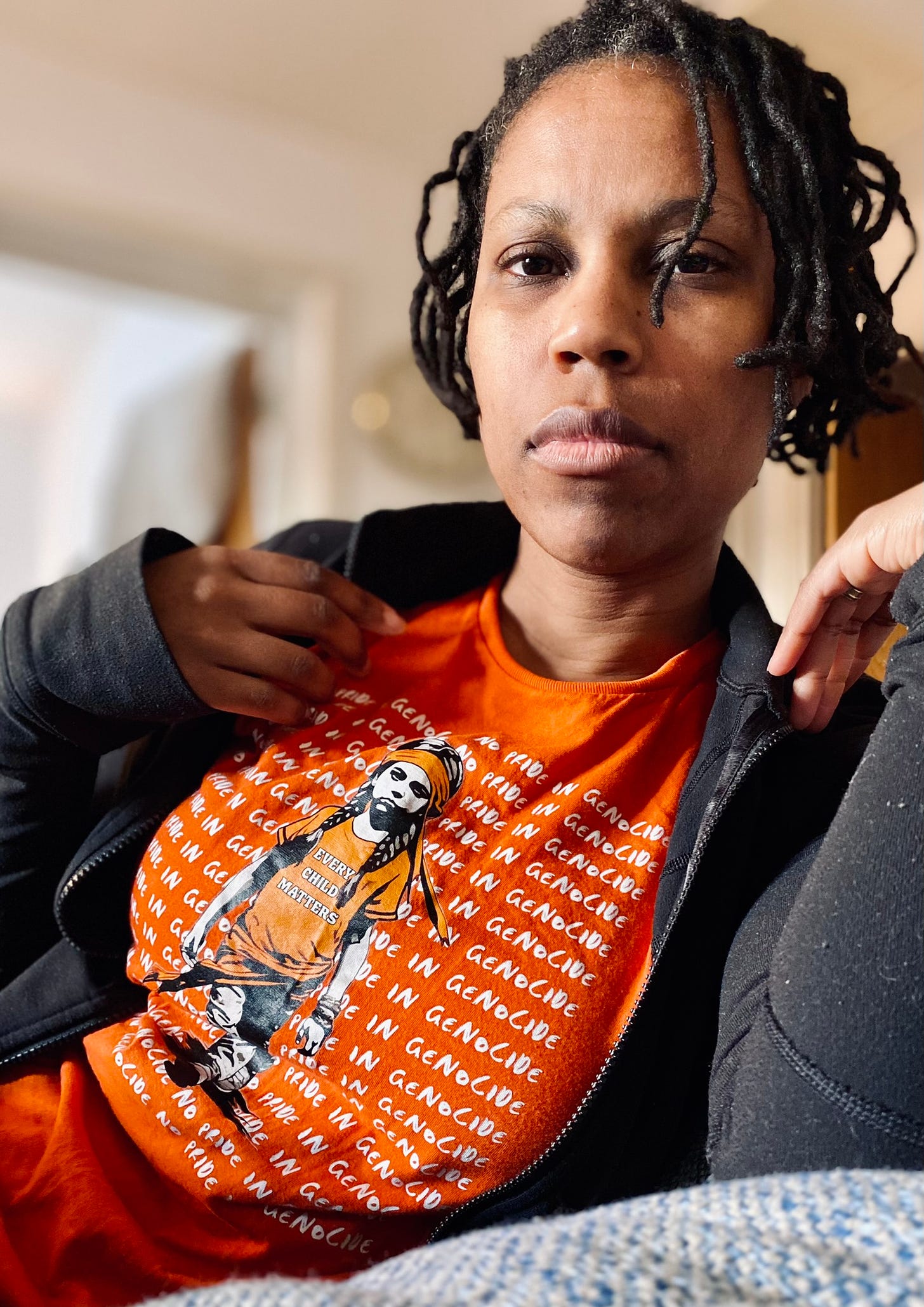 Sandra is a Black femme with short black locs staring into the camera wearing a bright orange shirt with an Indigenous child on it. The shirt repeats no pride in genocide. The Indigenous child wears a shirt that says, every child matters.