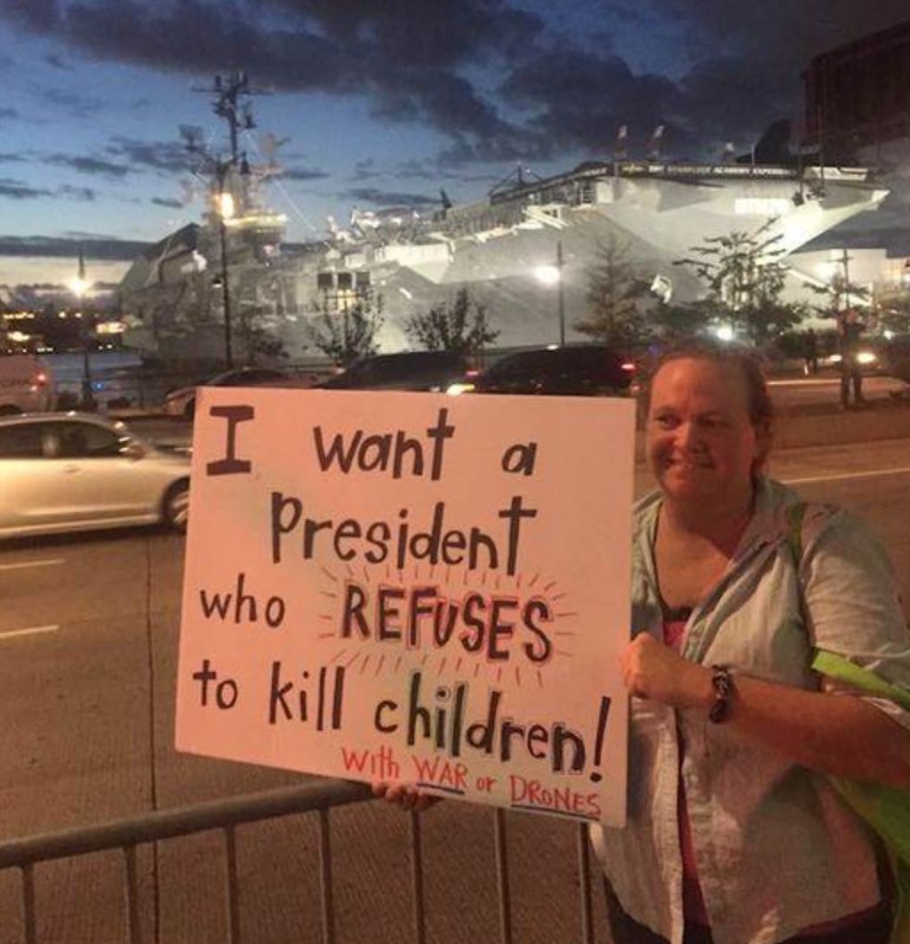 HePerson holding a sign that says "I want a president who refuses to kill children with war or drones, while standing in front of an aircraft carrier