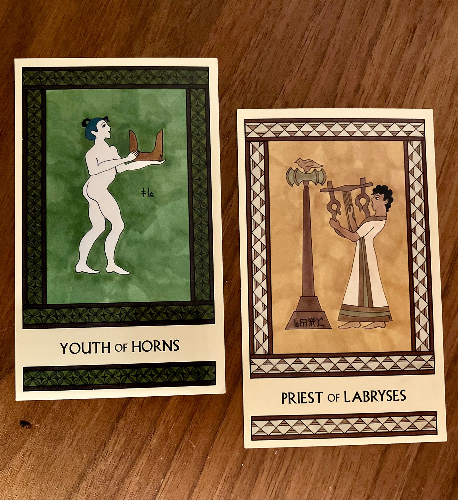 Two Minoan Tarot cards side by side on a wood surface. The Youth of Horns is in shades of green. It shows a young Minoan boy facing right, holding a pair of sacred horns. The Priest of Labryses is in shades of yellow and gold. It shows a Minoan man in priestly robes facing left, playing a lyre, with a tall labrys in front of him. 