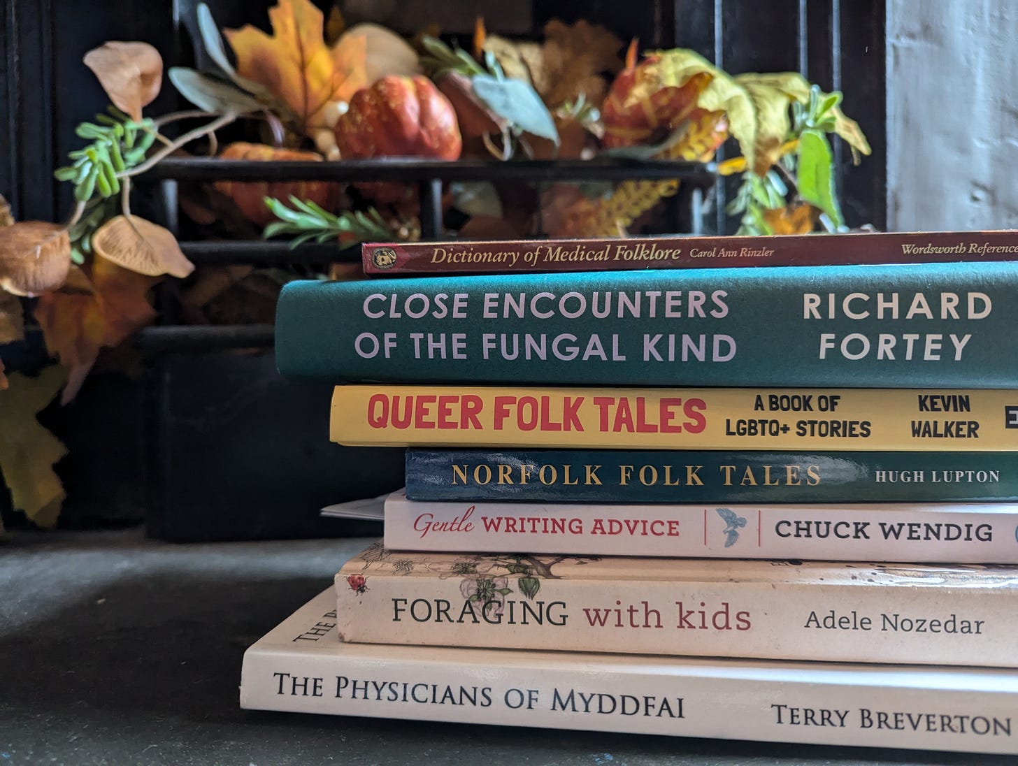 A pile of folklore, foraging, and writing books in front of some autumnal colours in a fireplace