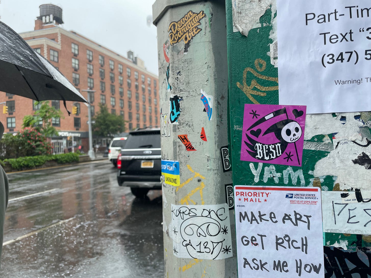 A label stuck to a telephone pole says in all-caps, printed with a Sharpie, “Make Art/Get Rich/Ask Me How”.