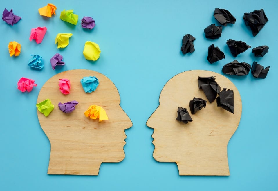 Two wooden outlines of human heads facing each other, the one on the left with colorful crumpled up PostIT notes rising from the brain, the other with all-black PostIT notes, indicating opposition to the creative ideas of the head on the left.