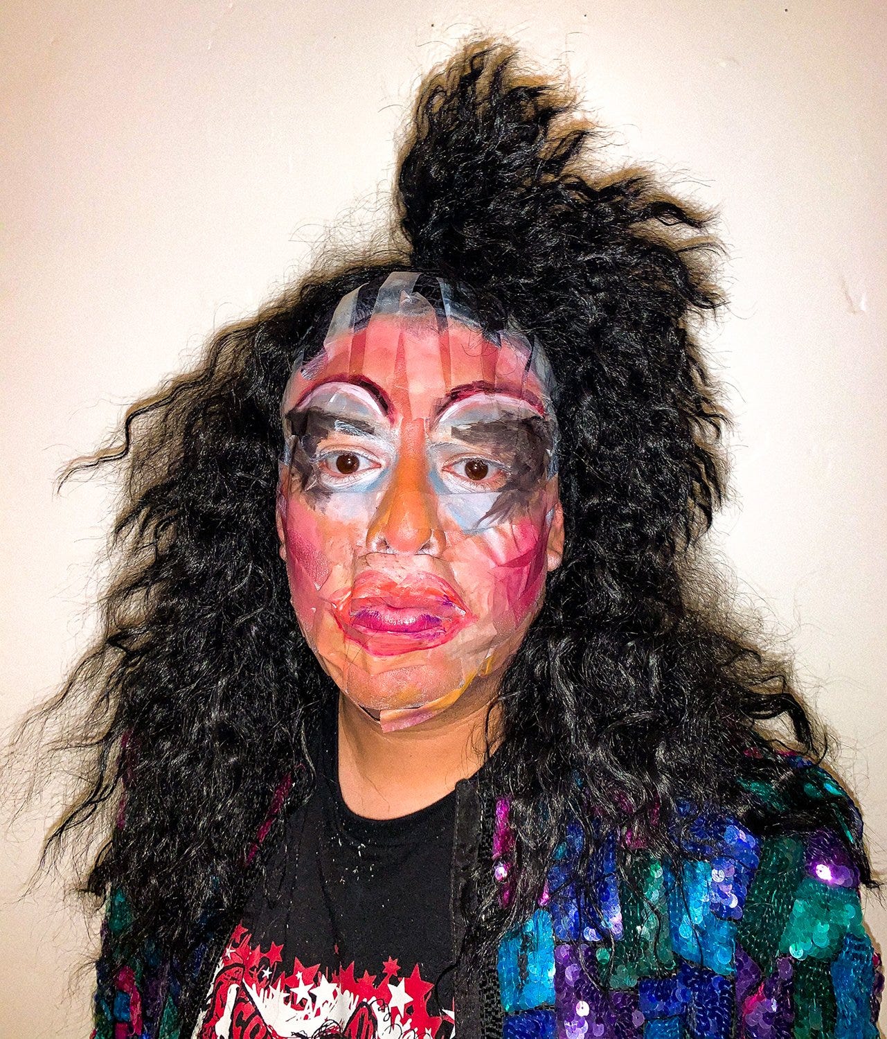 Photographic portrait of drag persona Krystal Lake Carrington in front of a white background. Her dark wavy hair is messy and teased in 80s big hair fashion and her makeup, including silvery-blue eyeshadow, magenta rouge, and fuschia lipstick, is very exaggerated and messy applied over strips of cellophane tape. Krystal Lake is wearing a black, magenta, teal, and electric blue sequined blazer over a black tee-shirt. Her face is serious and her gaze is deadpan, looking just down and off-camera. 