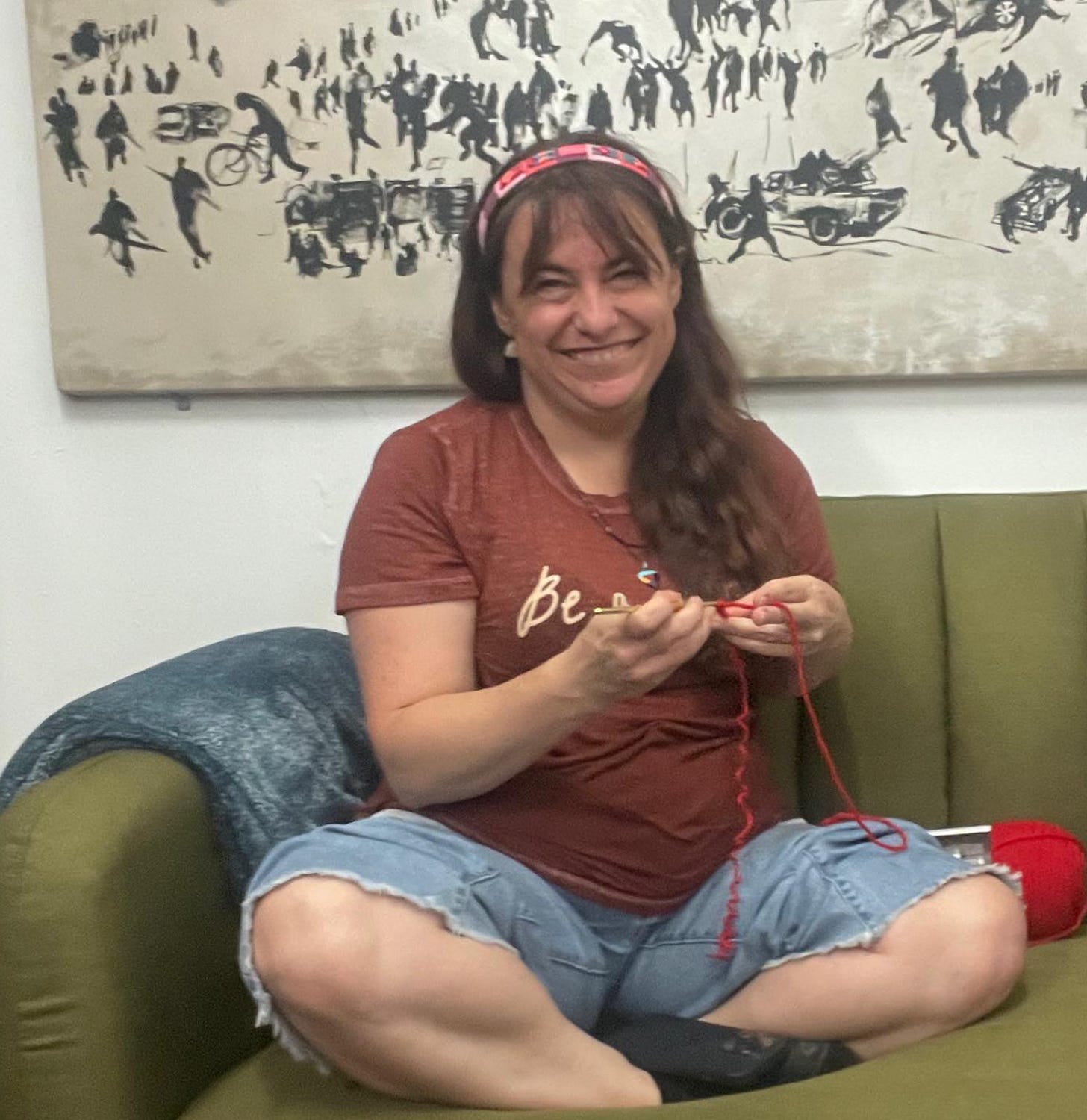 Kathryn sits, smiling, on a green couch, crocheting with red yarn