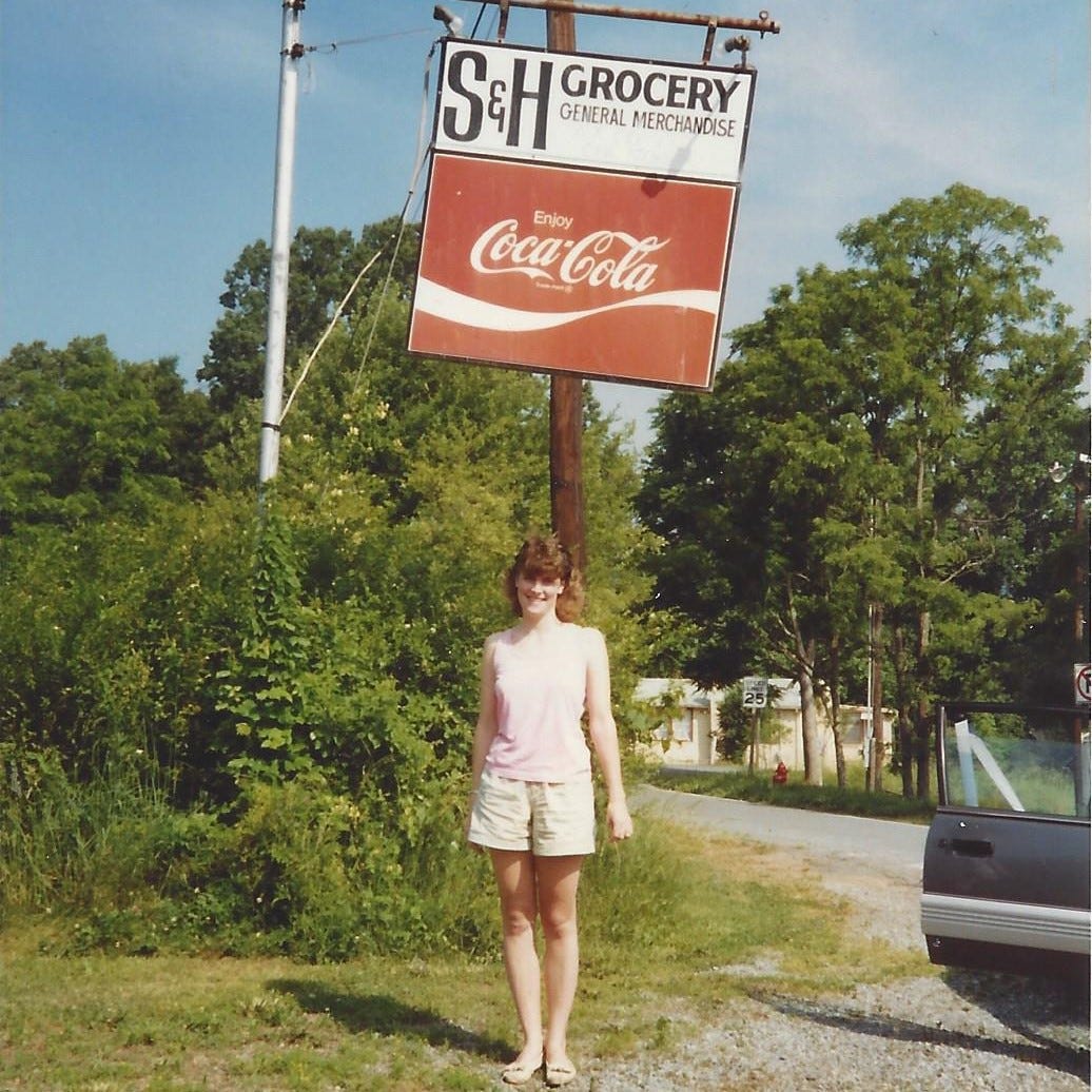 20-something woman standing under a Coca Cola sign for a small general store in the middle of nowhere