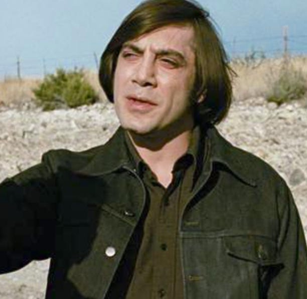 An image of Javier Bardem in a black denim jacket wearing maybe a dark wig with desert as backdrop.