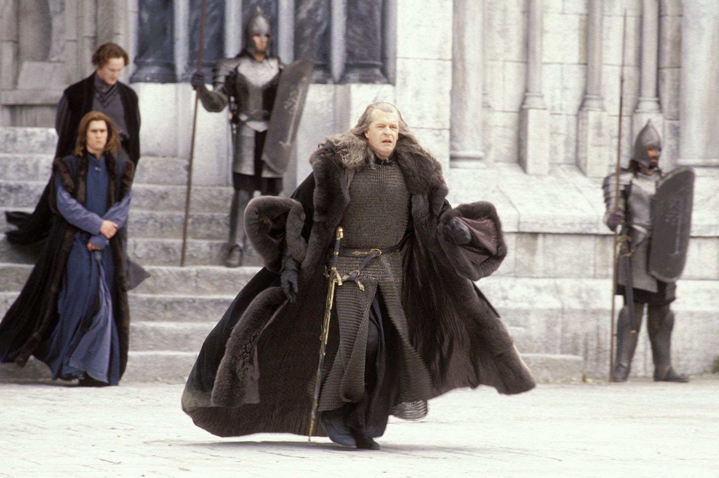 Denethor striding across a courtyard, his fur-hemmed velvet cloak billowing in the wind of his speed, revealing a full coat of chain maille and a sword at his hip.