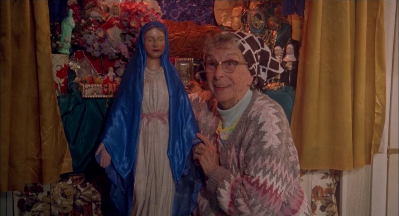 Image of an elderly woman in a sweater and headband, holding a statue of the Virgin and smiling, with a colourful display in the background. Image still taken from the movie "Pecker".