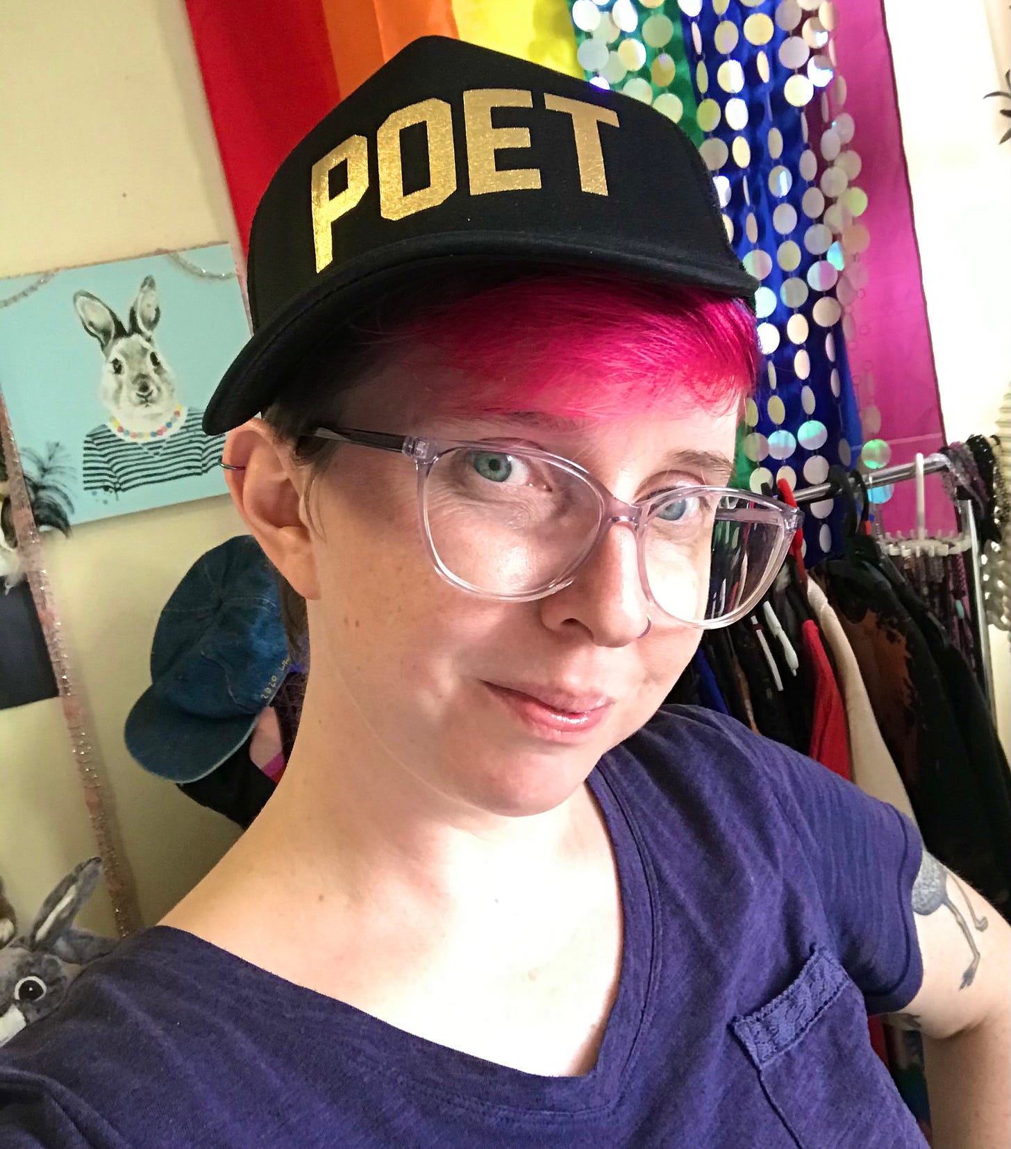 Rae White is in a room. They are wearing a cap with "poet" in gold on it. They have pink hair, large glasses and are wearing a purple shirt. in the room is a drawing of a rabbit.