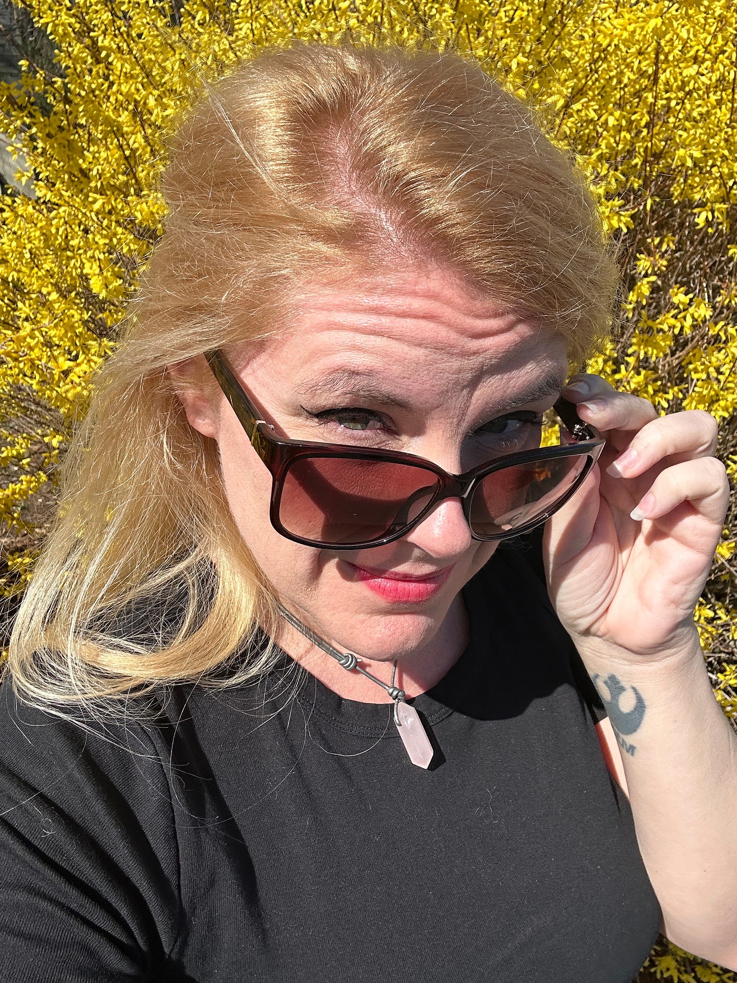 Cass in front of a field of forsythia, tipping her sunglasses down while looking at the camera