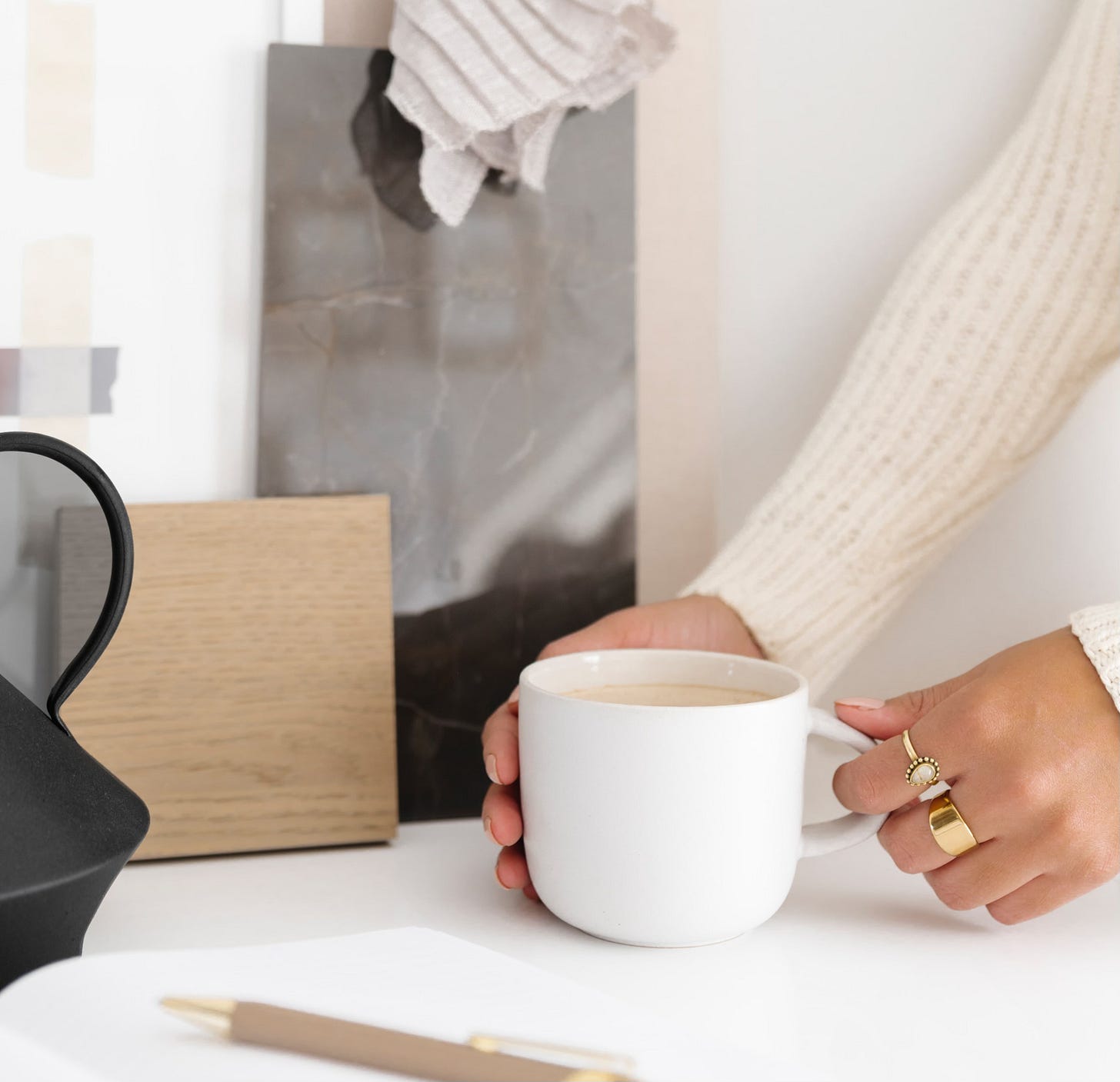 A person holds a white coffee mug at a bright desk with gold rings on their fingers. Nearby, a pen, notebook, and design swatches suggest quiet creative work.