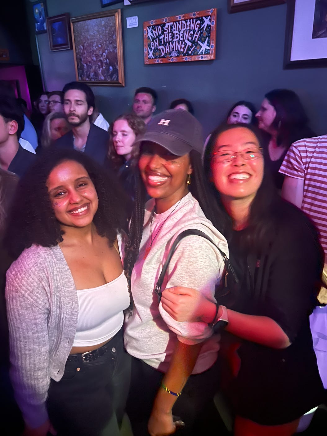 Three girls standing in a Jazz bar, crowd around them as they smile into the camera.