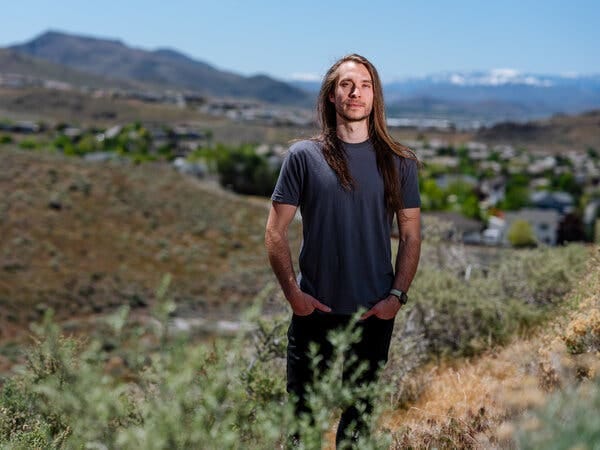 Jacob Sprague, with long brown hair past his shoulders and wearing a blue T-shirt and dark jeans, stands in an outdoor setting with his hands in both pockets.