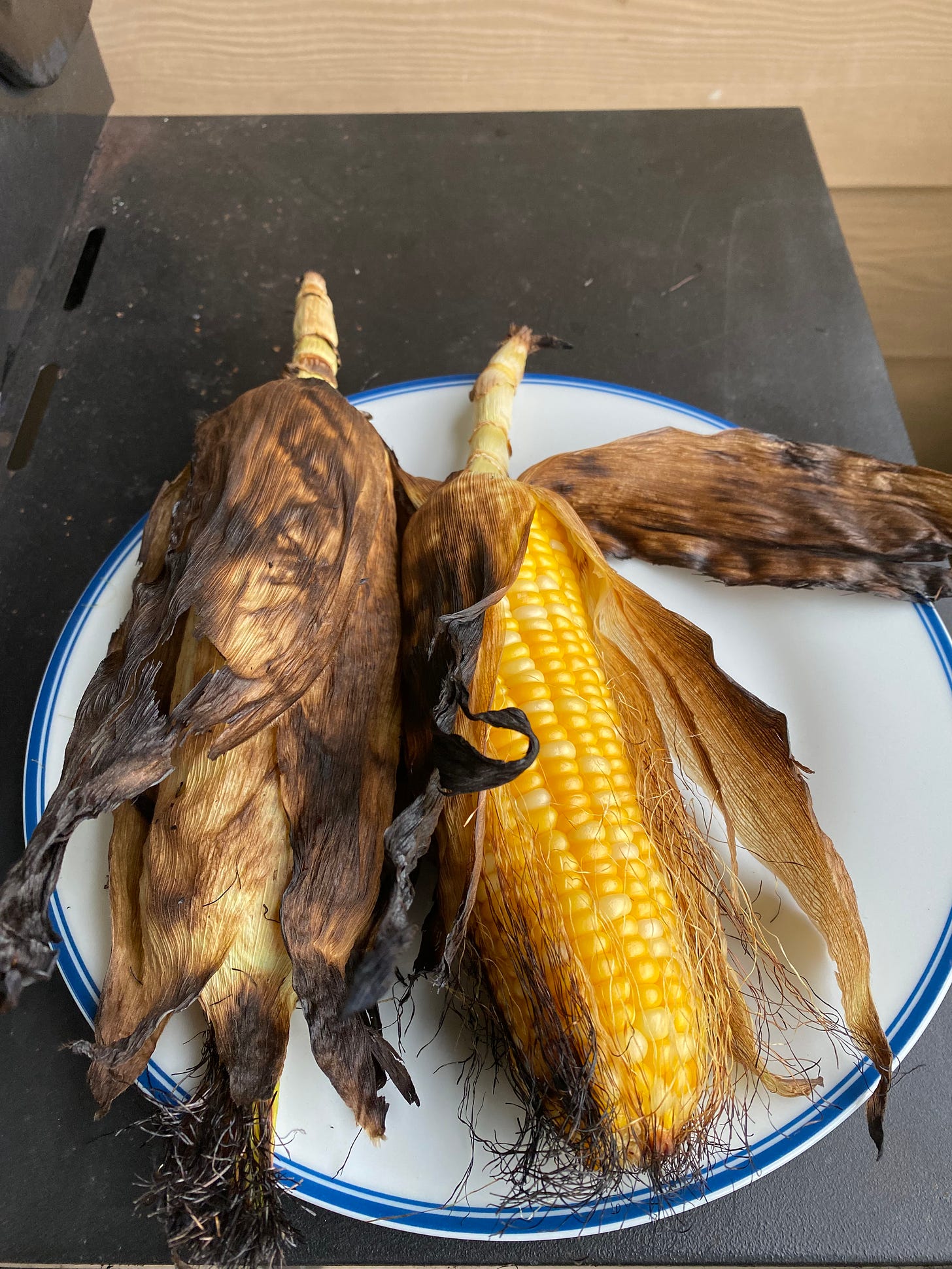 two grilled ears of corn on a plate beside a barbecue. One has the husk still mostly intact, but charred, and the other the husk has peeled mostly off, revealing peaches and cream corn beneath.